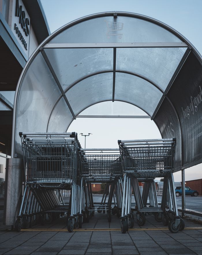 A group of metal shopping carts under a transparent shelter at a supermarket parking lot.